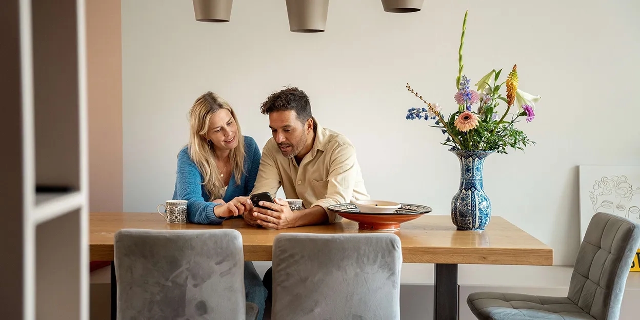 Man en vrouw zitten samen aan de eettafel en kijken op mobiele telefoon